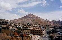 City of small simple houses in front of a large conical red mountain.