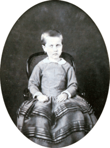 Photograph of a young Isabel with hair gathered at the back and wearing earrings seated on a small, cane back armchair