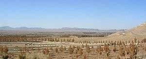 Sparse brown vegetation in dusty soil fills the foreground, fading to distant mountains along the horizon. A barely discernible scattering of buildings is in the middle distance.