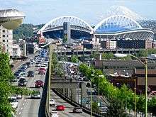 A stadium surrounded by historic buildings. A raised roadway runs along its side and a glacier capped mountain is behind it