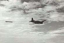 Black and white photo of a single-engined monoplane in flight. The aircraft is towing a piece of fabric.