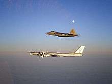 Aerial port view of two aircraft in flight, one on top of the other. The bottom aircraft is a four-engined propeller-driven aircraft, which is escorted by a jet fighter.