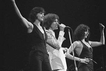 Three people sing onstage next to each other. The man in the middle wears white clothes, and on both sides stands a woman wearing black.