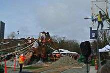 An action shot of people, wearing orange and yellow construction clothing, working from the ground and from a crane during the raising of the Reconciliation Pole at UBC.