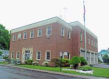 A roughly cubical two-story brick and concrete building with metal lettering spelling out "Village of Rhinbeck" above the entrance. On the left side are four garage doors and a flagpole from which an American flag hangs.