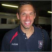 A man in a navy blue open-necked shirt with a crest and the word "mitre" on the chest, smiling.