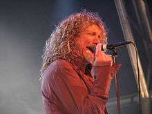 A man with long, curly hair wearing a red dress shirt and singing into a microphone on a stand.