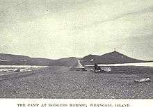 A conical tent stands on a desolate stretch of beach, with a range of low hills in the background.