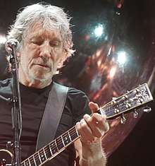 Roger Waters playing a bass guitar and singing into a microphone. He has grey hair and is unshaven.