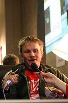 Man with short, wind-swept, dirty-blonde hair speaking into two microphones