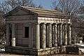 Light colored stone mausoleum flanked by four columns. Door is aged metal.