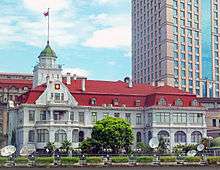 An ornate stone building with a red roof. From a cupola on the left side the Russian flag is flying. There is a taller skyscraper behind the right side.