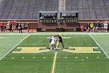 Two women, slightly hunched forward to each other, one in a black sleeveless top and short skirt and the other in a white version of the same clothing, stand in the middle of an artificial turf field with markings for gridiron football, while a third woman wearing a vertically black and white striped shirt stands just behind them with one hand between them, seen from some distance and above