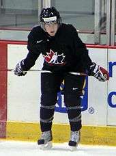 A Caucasian teenage ice hockey player. He stands crouched over towards the ice with his stick resting laterally on his thighs. He wears a black jersey with a stylized maple leaf logo.