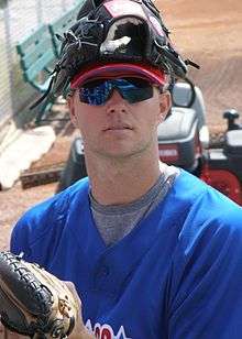 A man in a blue baseball jersey and wearing a catcher's mitt on his left hand squats in a bullpen. He has a catcher's mask made of leather and metal atop his head.