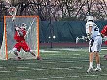 A man wearing a red jersey and shorts with a white helmet and gloves holds a white stick with a large meshed area on one end just above a square of metal roads with netting behind it. His pose, with his legs apart, left knee bent and back curved slightly, suggests great effort in doing so. On the right another man, wearing white jersey and green trim but otherwise similarly attired to the first, holds a similar stick with a much smaller head at his right. Behind the scene is a wall covered with black tarpaulins, and trees against an orangey sky