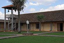 A church courtyard with a sundial in the center of the courtyard and a wooden bell-tower in the corner of the courtyard. The bell tower consists of a roofed platform supported by four wooden columns. Stairs lead up to the platform. The sundial is located at the end of a wooden column.