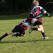 A child running away from camera in green and black hooped rugby jersey is in the process of being tackled around the hips and legs by an opponent.