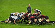 Two opposing formations of eight men, in white and black to the left, red and black to the right, push against each other in a crouched position; behind them stands another player and the referee.