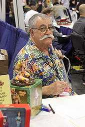 A man with thinning, greying hair and a thick, white mustache sits behind a table. He wears a colourful shirt. Pens and action figures of his works are on the table in front of him.