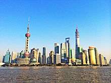 A group of skyscrapers and towers, seen from across a river. At the left is one consisting of a sphere on concrete supports topped by a long spike; in the center are smaller buildings, one a bright gold color, gradually rising to the tallest one at right, still under construction