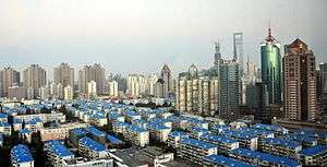 A cityscape seen from a height. In the foreground are low-rise buildings with blue roofs, giving way at the rear to higher-rising towers of different shapes and materials