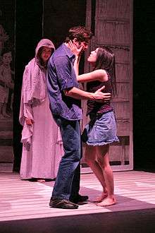 A photograph of a man wearing a blue dress shirt and blue jeans and looking into the eyes of a woman wearing a brown shirt and a blue jean skirt