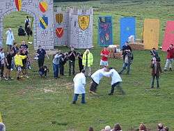 Two men, both in white shirts and jeans, are grasping each other at the shoulders and attempting to kick each other. They are surrounded by an onlooking crowd