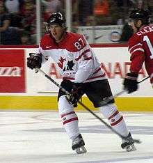 Hockey player in red and white uniform, with a maple leaf picture on his chest. He skates on the ice, looking beyond the camera.