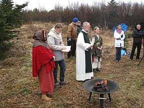 Eight humans, all white, stand on heathland. A number of them are dressed in historical clothing akin to that worn in the medieval period.