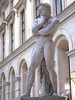 A marble statue of Spartacus in front of a neoclassical building. He is standing naked with his legs astride, looking into the middle distance with his left hand clenched against his chin, his right arm crossing his chest on which his left arm rests.