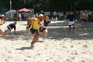 On a sunlit beach two teams of players, one in yellow the other in blue, play a form of rugby; the central yellow player runs forward clutching the ball with one hand, close to his chest.