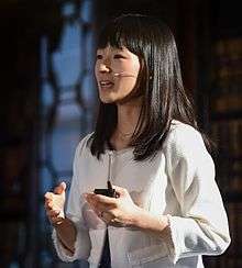 Waist high portrait of woman with long dark hair and bangs wearing white jacket speaking into a microphone