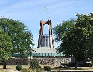 A modern church building with a green, domed roof, a glass tower above the roof, and brown arches above the tower