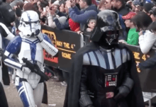 Two costumed soldiers, one in white and purple armor and the other in black, parade in front of fans taking photographs.