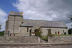 Stone building with small square tower. In the foreground is a road and wall.