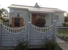 A small, detached one-story house. The walls are a pale colour, and the roof is made from corrugated iron.