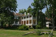 A large stone house with white dormers, window shutters and porch railings, surrounded by trees and shrubs.