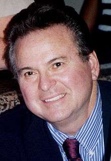 A man with dark hair, wearing a suit, including a purple tie and blue and white shirt.