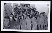 A grouping of about 30 students in front of a brick building. There is a nun in the back row.