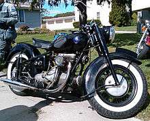 A mildly customized Sunbeam S7 motorcycle sits along the edge of a driveway.  It shows evidence of normal wear and tear, but appears to be in very good condition.  It is all black, with a vaguely eggplant-shaped fuel tank that features rubber pads for the rider's knees, and white-wall tires.