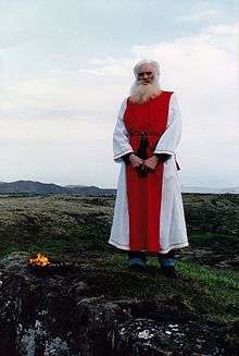 An elderly man wearing red and white robes standing in an open area.