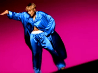 A women wearing a barely-buttoned and wind-blowing bright blue silk pajamas, showing a white boxer inside her pants, with her right hand up and her left hand on her hip, she is backed by a dark pink background.