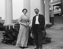 A man posing in front of several large Greek columns. He is about sixty-years-old and is wearing a two piece black suit and bow tie with a hand in his pocket and holding a wide brimmed white hat in the other while standing in a casual pose with a partial smile and his hair being blown up by the wind. A woman stands to his right wearing a full length gown and holding a folded fan between her clasped hands; her glasses obscure her eyes and while she stands in a dignified pose.