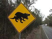 A square-shaped metal tilted at 45 degrees on a metal post. The sign is painted yellow with a picture of a black devil in profile. It is at the side of a straight road cutting through wooded forest and two vehicles can be seen.