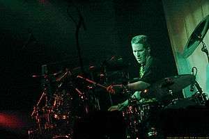 Drummer with crewcut, wearing black T-shirt, intently plays his large drum set in the spotlight.