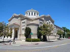 The corner of a two-story brick building faces the intersection of two streets. To the right, the building presents many small windows, with three taller ones near the corner, all framed in white. To the left, the building presents more small windows framed in white, and a portico supported by multiple white columns. Atop the building is an elliptical dome which has white-framed windows on its sides.