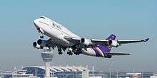 An airborne Boeing 747 passing in front of an airport terminal and control tower, which are blurred behind the exhaust from the plane's jet engines. The plane's flaps are extended, and its landing gear still deployed.