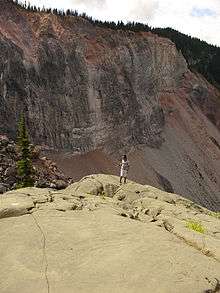 A large rocky cliff rising above rubble with a person in the foreground standing on a rounded rock.