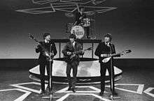 Monochrome image of The Beatles performing on a stage wearing dark suits